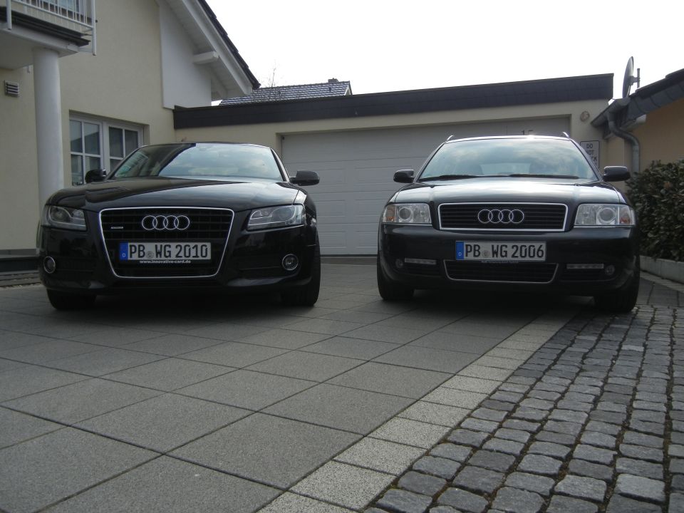 Two black Audi sedans parked in a driveway. The car on the left has license plate PB WG 2010. The car on the right has license plate PB WG 2006