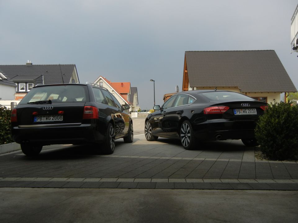 Two black Audi cars parked in a driveway. The one on the left is a wagon with license plate PB WG 2006. The one on the right is a sedan with license plate PB WG 2010