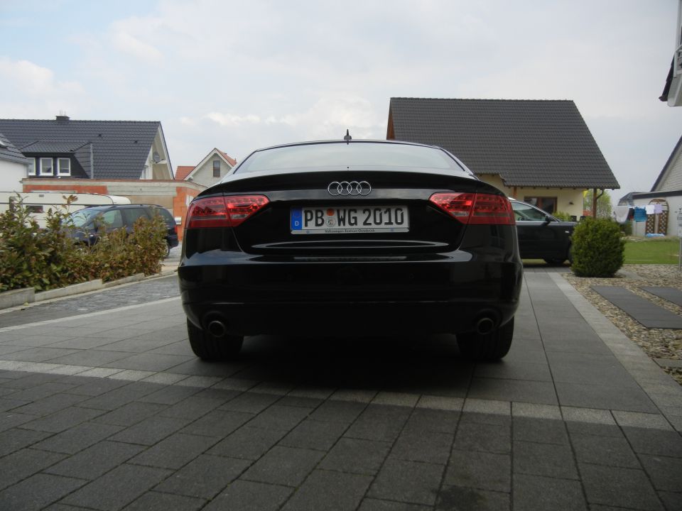 Rear view of a black Audi with license plate PB WG 2010 parked in a driveway. Another car is visible in the background