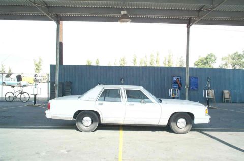 1991 Ford LTD Crown Victoria - A white sedan is parked under a metal awning, with a blue fence in the background.