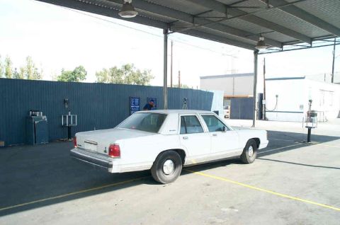 1991 Ford LTD Crown Victoria - A white sedan is parked outdoors under a metal roof, with a blue wall and buildings in the background.