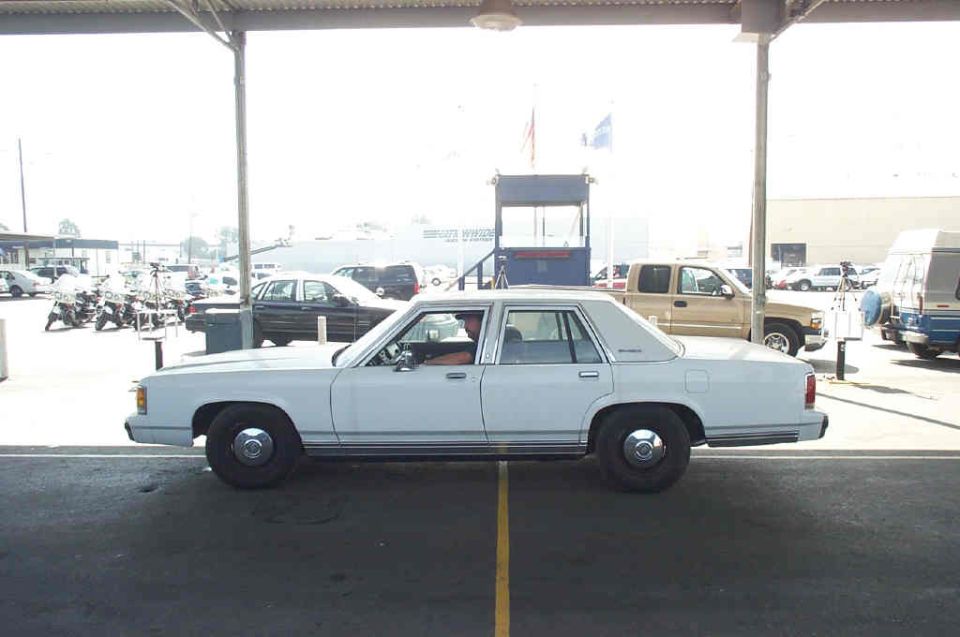 1991 Ford Crown Victoria LTD sedan parked under a structure, side view. Other vehicles and a guard booth are visible in the background