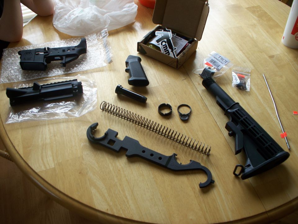 Gun parts laid out on a wooden table, including a receiver, stock, spring, and tool