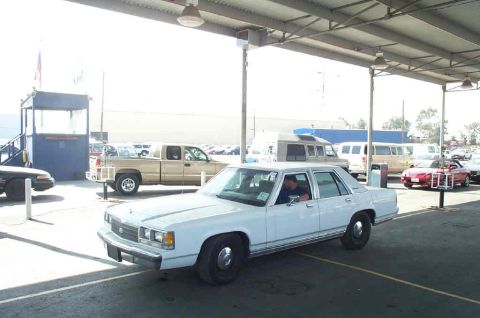 1991 Ford LTD Crown Victoria - A white sedan is parked under a canopy, with a driver visible inside. Several other vehicles are in the background.