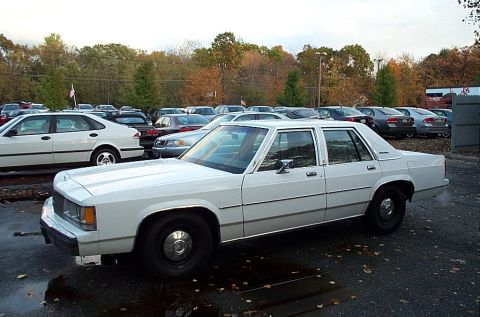 1991 Ford LTD Crown Victoria - This white sedan, likely from the 1980s, is parked in a lot with other vehicles under an overcast sky.