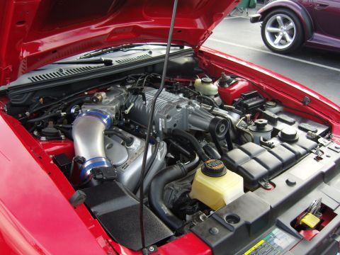2007 Ford Mustang - Close-up of a red vehicle's engine compartment, a prominent supercharger and intake system. Another car is visible in the background.