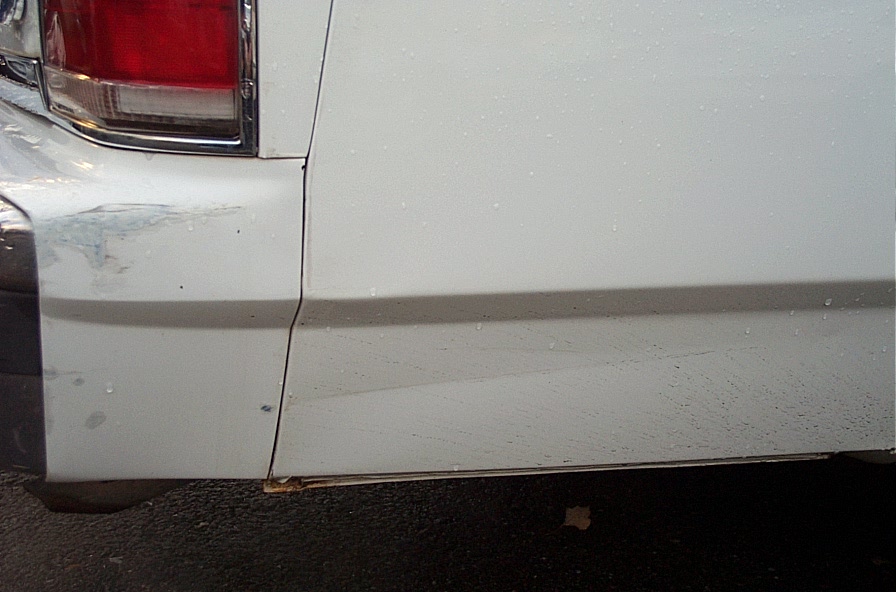 Close-up of white car's rear fender and bumper area, showing some scuffs and water droplets
