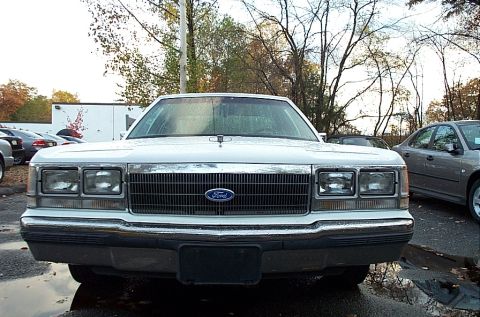1991 Ford LTD Crown Victoria - A white 1991 Ford Crown Victoria LTD is parked outside on a wet day, showing its front grille and headlights.