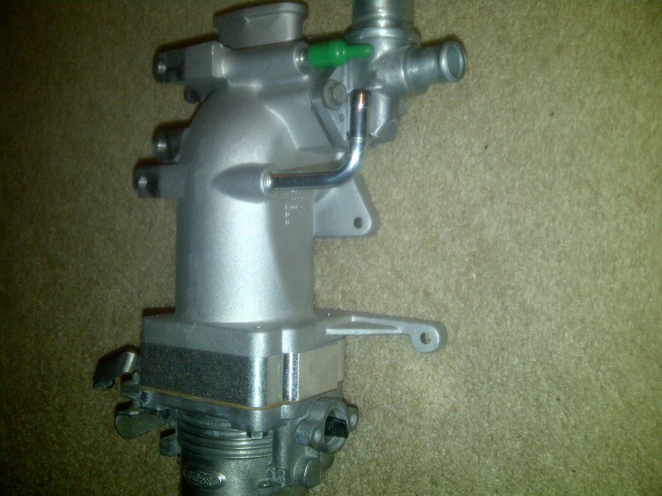 Silver throttle body with green valve and metal tubing, resting on carpet