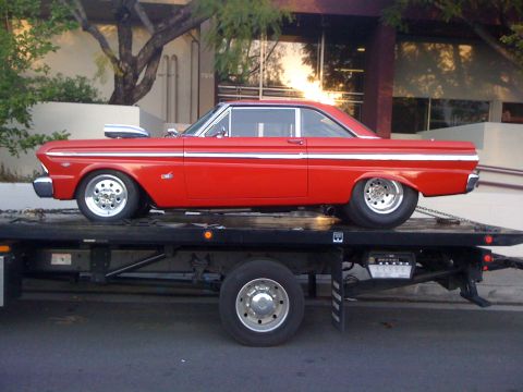 1964 Ford Ranchero - A red 1964 Ford Falcon coupe is loaded onto a tow truck, its chrome trim and hood scoop visible.