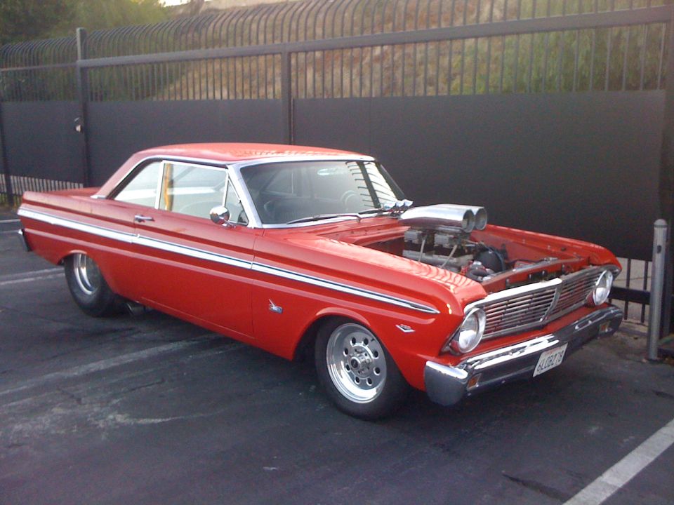 Red 1964 Ford Falcon coupe with a chrome supercharger protruding from the hood, parked in front of a black fence