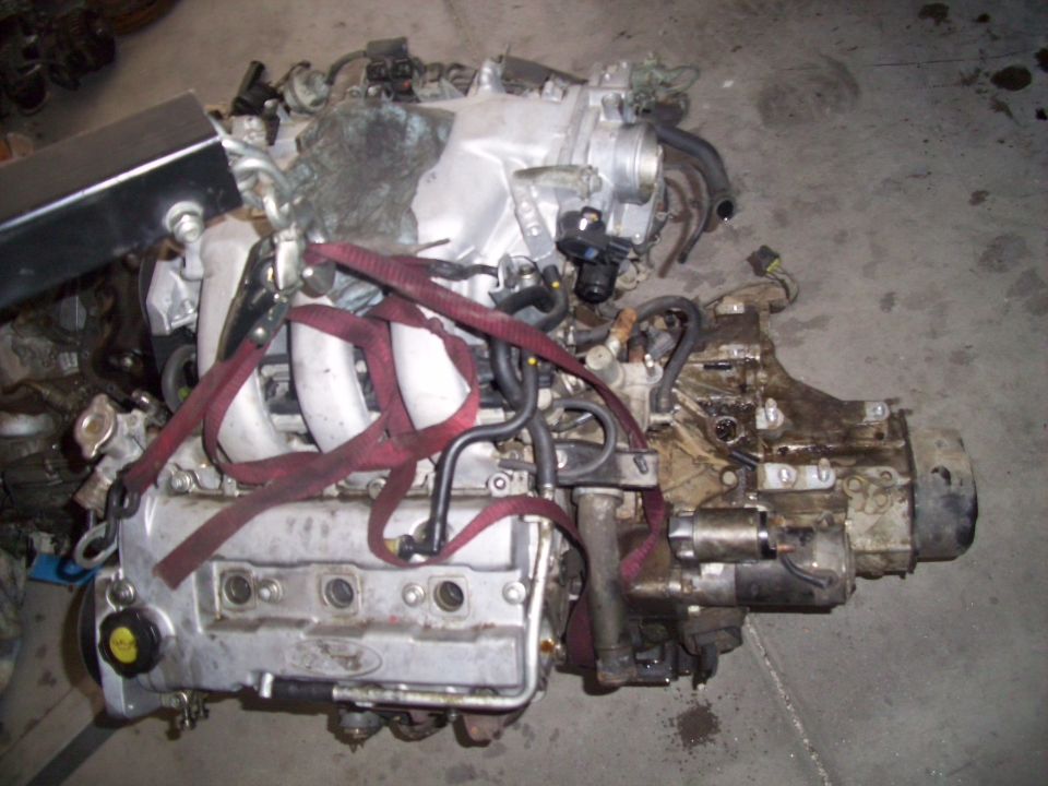 Engine with red straps, silver valve cover with Ford logo, and transmission attached