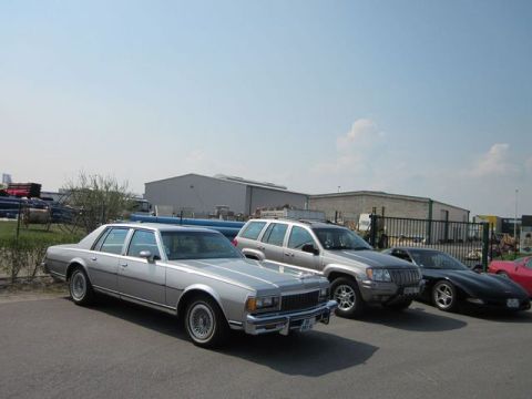 1991 Ford LTD Crown Victoria - Several vehicles are parked in front of industrial buildings under a clear sky.