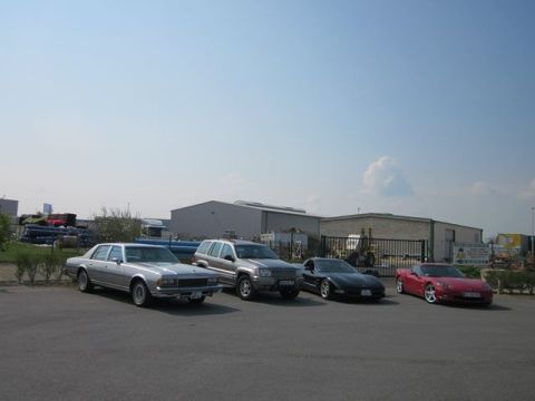 1991 Ford LTD Crown Victoria - A collection of vehicles, including a vintage sedan and modern sports cars, are parked outside industrial buildings under a clear sky.