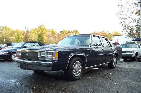 1991 Ford LTD Crown Victoria - Dark blue Ford LTD Crown Victoria four-door sedan parked on wet asphalt among other cars, photographed from the 3/4 front left with autumn…