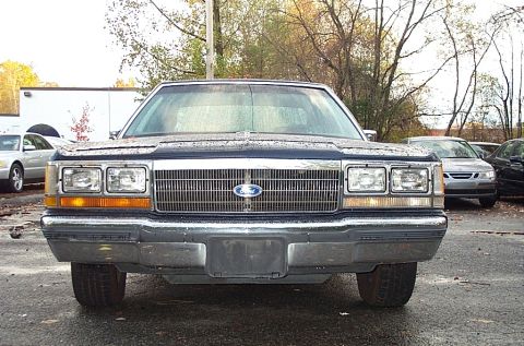 1991 Ford LTD Crown Victoria - A dark blue Ford Crown Victoria is parked on a wet lot, showing its front grille and headlights.