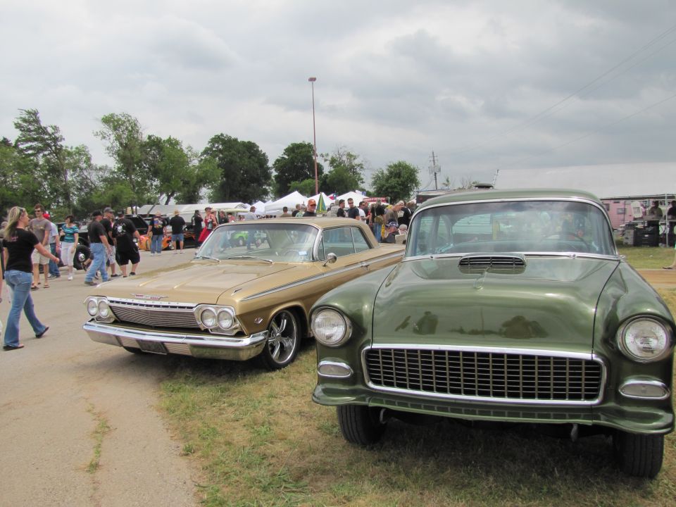 Gold 1962 Chevrolet Impala parked next to a green 1955 Chevrolet. People and tents in background