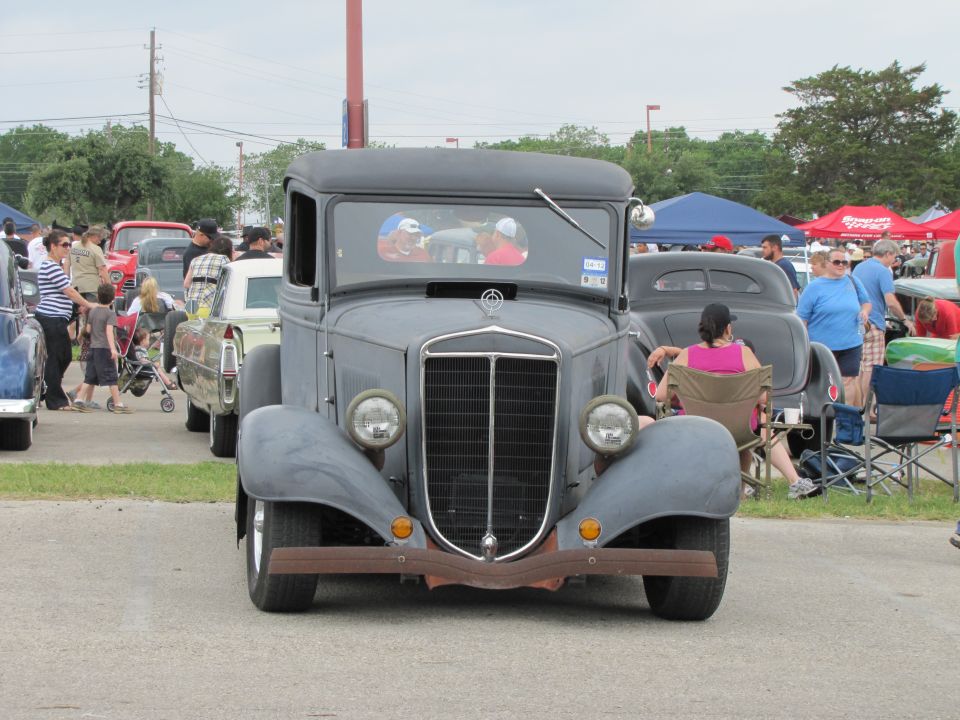 Gray vintage pickup truck with a prominent grille and round headlights. People and other cars are visible in the background