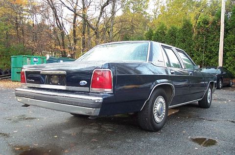 1991 Ford LTD Crown Victoria - A dark blue Ford sedan is parked in a lot with trees and dumpsters in the background.