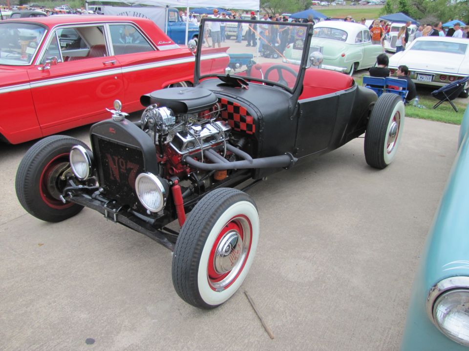 Front view of a black and red hot rod with a large engine and whitewall tires. A red classic car is to its left