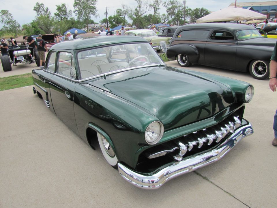Green customized 1950s sedan with wide whitewall tires and chrome grille, parked at an outdoor car show. Another dark panel van is behind it