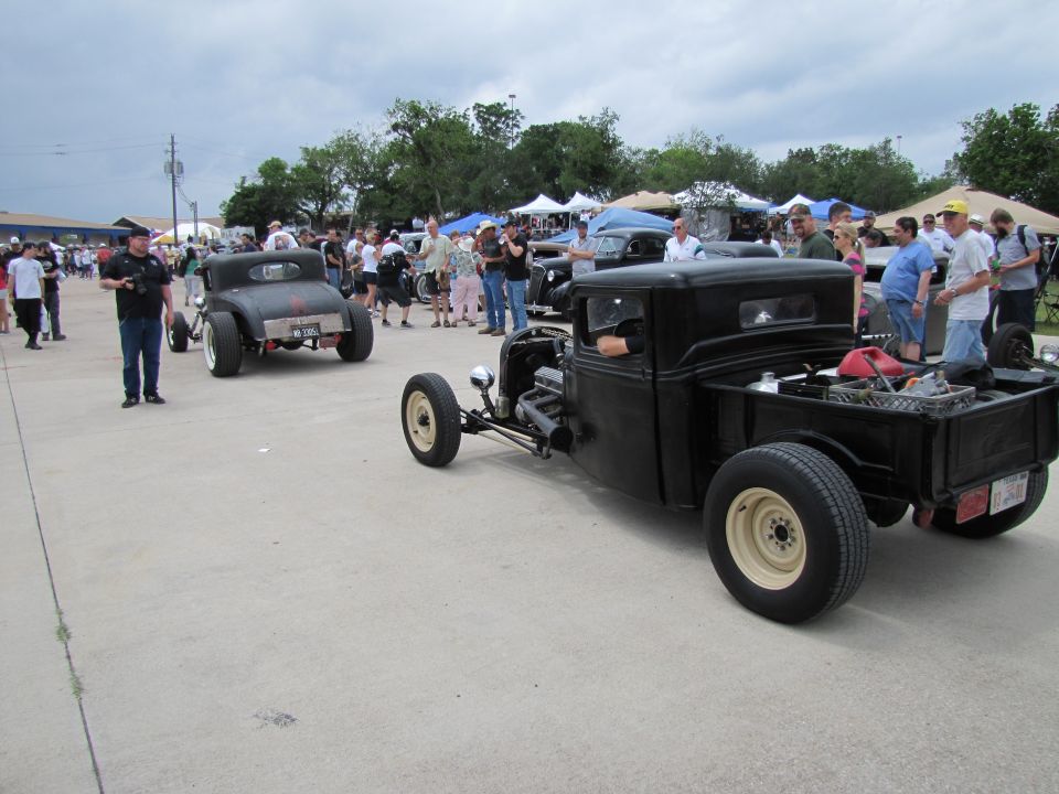 Black hot rod pickup truck in foreground, another hot rod car behind it, people milling around tents in background