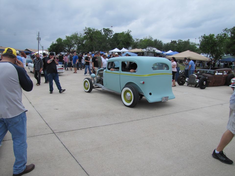 Light blue hot rod sedan with yellow trim, white wall tires, and exposed engine components, viewed from the rear