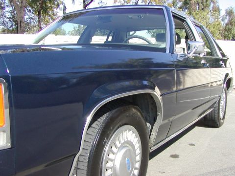 1991 Ford LTD Crown Victoria - A dark blue sedan is parked on an asphalt surface, with trees and a wall in the background.