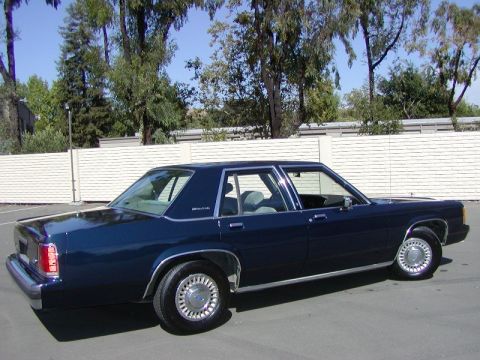 1991 Ford LTD Crown Victoria - A dark blue sedan is shown from the side, parked outdoors with trees and a white fence in the background.