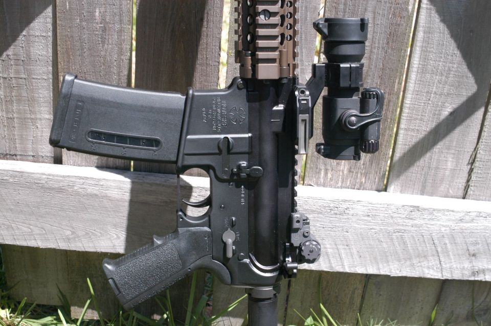 AR-15 rifle with magazine and optic mounted, resting against a wooden fence