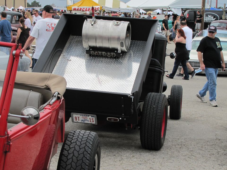 Rat rod truck with a keg in the bed, next to a red convertible. People walking around