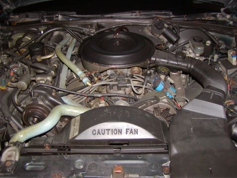 1991 Ford LTD Crown Victoria - Engine bay of an older Ford V8 shot from the front, with a round black air cleaner over the throttle body, a light-green upper radiator hos…