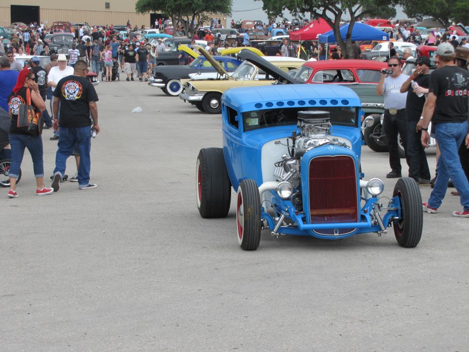 blue hot rod with exposed engine and red grille, parked at an outdoor car show with other vehicles and people