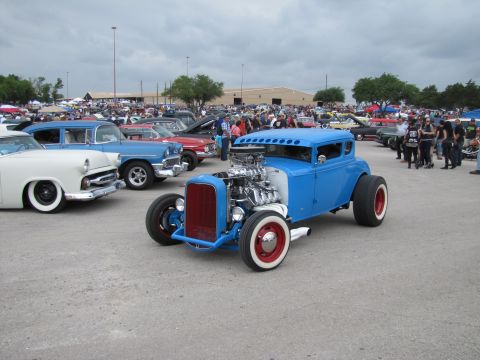 2006 BMW 5 Series - A crowd gathers around various classic cars at an outdoor event, with a prominent blue hot rod in the foreground.