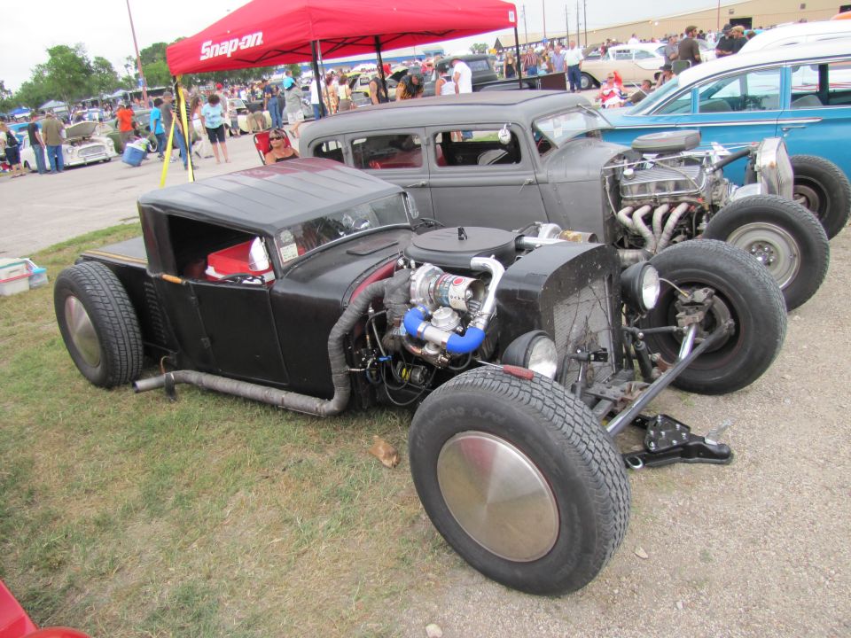 Custom hot rod with exposed V8 engine and turbocharger, parked next to a grey sedan and a blue car