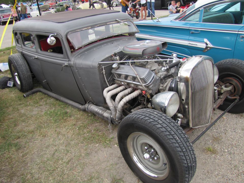 Custom hot rod sedan with exposed engine and side pipes, parked next to a blue car