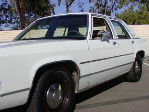1991 Ford LTD Crown Victoria - A white sedan is parked outdoors, with a brick wall and trees in the background.