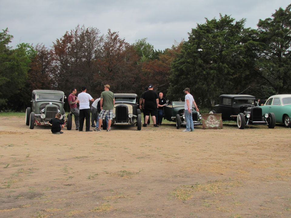 Several hot rod cars parked on dirt with people standing around them