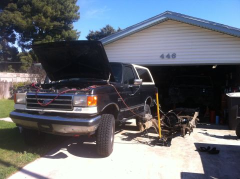 1989 Ford Bronco - A Ford Bronco is being worked on outside a garage, with its chassis detached and another vehicle inside.