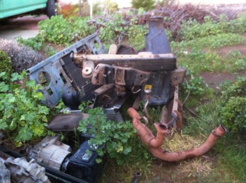 1989 Ford Bronco - Disassembled vehicle components, including a frame and engine parts, are scattered on the ground amidst overgrown vegetation.