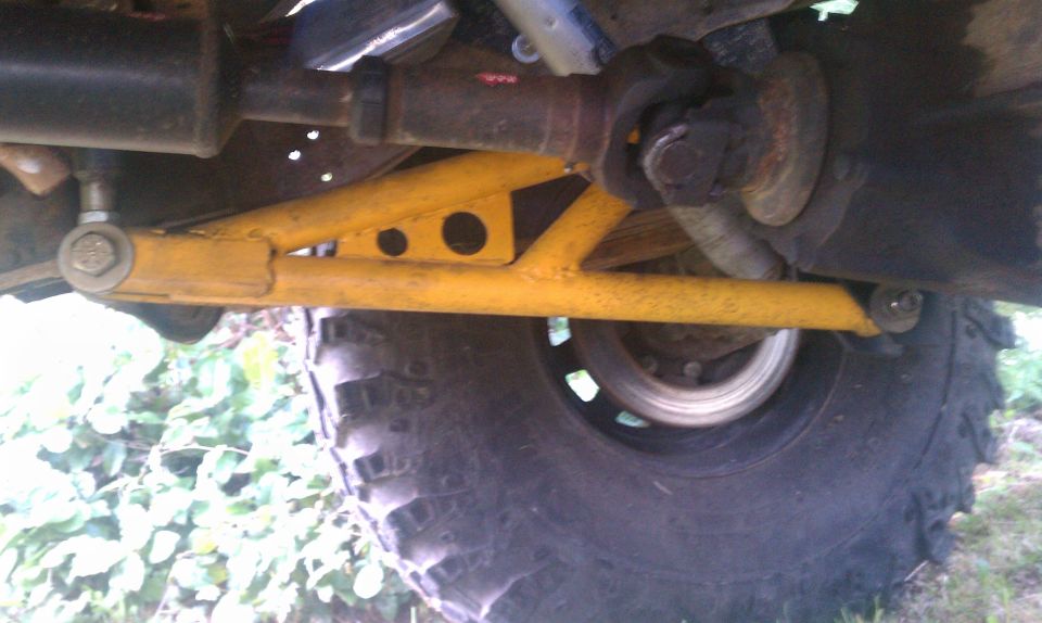 Yellow suspension arm and driveshaft visible beneath a large tire and wheel. Some green foliage is in the background