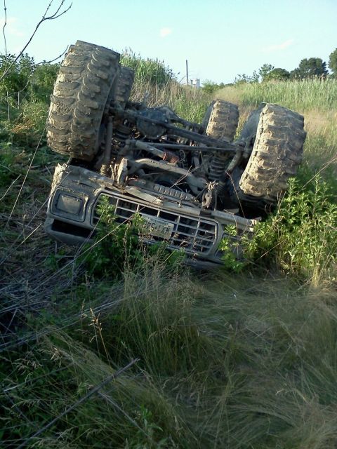 1990 Ford Bronco - An overturned off-road vehicle rests on its side in a grassy field under a clear sky.