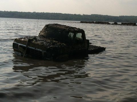1990 Ford Bronco - Mud-covered truck partially submerged in water, with trees and a breakwater in the background.