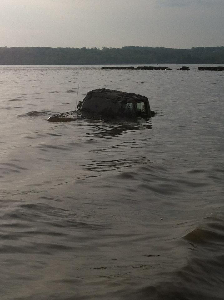 Mud-covered SUV is partially submerged in water, with only the roof and part of the sides visible above the surface