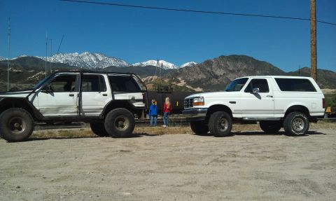 1978 Ford Bronco - A white Ford Bronco is parked next to a modified SUV, with a train car and mountains visible in the distance.