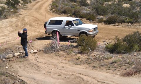 1978 Ford Bronco - A person photographs a white Ford Bronco navigating a dusty, off-road trail with sparse desert vegetation.
