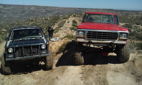 1978 Ford Bronco - Two modified off-road vehicles navigate a challenging dirt path in a mountainous, arid landscape.