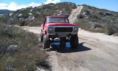 1978 Ford Bronco - This lifted pickup truck is navigating a rough dirt path in a dry, hilly area under a clear blue sky.