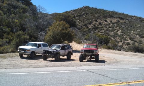1978 Ford Bronco - Three modified off-road vehicles are parked on a dirt pull-off next to a paved road with a mountainous, scrub-covered hillside in the backg…