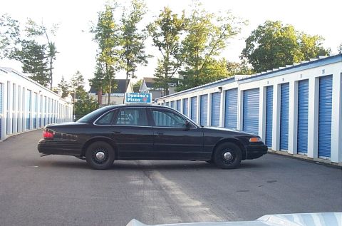 2001 Ford Crown Victoria - A black 2001 Ford Crown Victoria, possibly a former police car, is parked outside storage units with a Domino's Pizza sign on its roof.